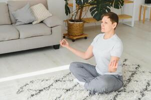 Working with your body. Full length of beautiful middle aged woman in sportswear doing pose lotus while sitinding in front of window at home. photo