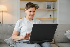 Elderly female sit on couch holding on lap laptop search information, check or typing e-mail, spend time on-line, modern tech, easy interesting pastimes services for retired older generation concept. photo