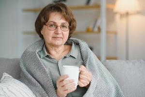 elderly caucasian pensioner wrapped in gray plaid and drinking hot tea in living room photo