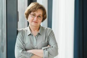 Portrait of beautiful senior woman. Elderly lady is standing by window at home. Active longevity concepts photo