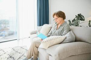 sosegado lectura. retrato de pensativo Envejecido mujer leyendo favorito literatura a acogedor hogar. ella es acostado en almohadas en cómodo sofá. piernas son cubierto con suave frazada. foto