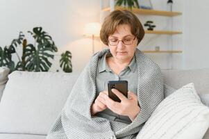 bella anciana con ropa informal usando su teléfono inteligente mientras se sienta en el sofá de casa foto