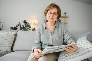 mayor dama leyendo su periódico a hogar relajante en un sofá y mirando terminado el parte superior a el espectador foto