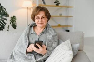 bella anciana con ropa informal usando su teléfono inteligente mientras se sienta en el sofá de casa foto