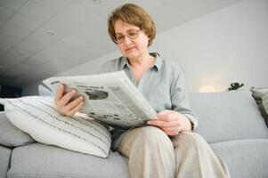 mayor dama leyendo su periódico a hogar relajante en un sofá y mirando terminado el parte superior a el espectador foto