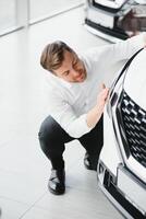 Happy handsome bearded man buying a car in dealership, guy hugging hood of new car photo
