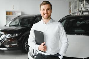 portrait of open-minded professional salesman in cars showroom, caucasian man in white formal shirt stands next to luxurious car and looks at camera photo