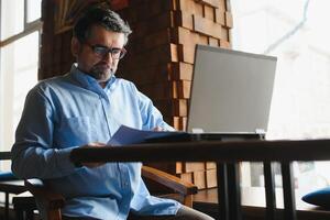 Male freelancer is working in a cafe on a new business project. Sits at a large window at the table. Looks at a laptop screen with a cup of coffee photo