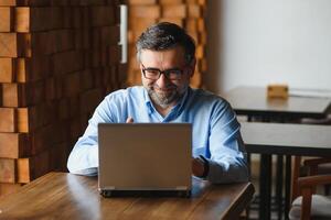 Male freelancer is working in a cafe on a new business project. Sits at a large window at the table. Looks at a laptop screen with a cup of coffee photo