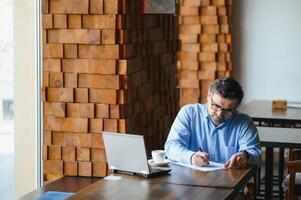 Male freelancer is working in a cafe on a new business project. Sits at a large window at the table. Looks at a laptop screen with a cup of coffee photo