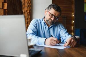 Positive senior bearded man with grey hair drinking coffee and using laptop at cafe, copy space. Stylish aged businessman in burgundy jacket enjoying his tea while working online. photo