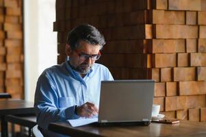 Male freelancer is working in a cafe on a new business project. Sits at a large window at the table. Looks at a laptop screen with a cup of coffee photo