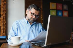 Male freelancer is working in a cafe on a new business project. Sits at a large window at the table. Looks at a laptop screen with a cup of coffee photo