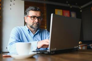 Male freelancer is working in a cafe on a new business project. Sits at a large window at the table. Looks at a laptop screen with a cup of coffee photo