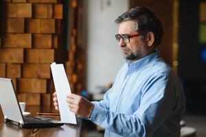 Mature businessman drinking coffee in cafe. Portrait of handsome man wearing stylish eyeglasses using laptop, looking at camera, smiling. Coffee break concept photo