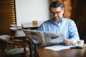 de cerca retrato de grave mayor hermoso hombre leyendo periódico, teniendo café descanso y sentado a mesa. foto