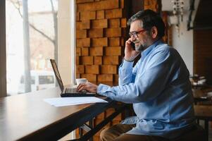 Positive senior bearded man with grey hair drinking coffee and using laptop at cafe, copy space. Stylish aged businessman in burgundy jacket enjoying his tea while working online. photo