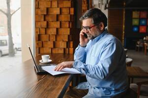 Male freelancer is working in a cafe on a new business project. Sits at a large window at the table. Looks at a laptop screen with a cup of coffee photo