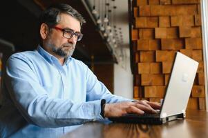 Male freelancer is working in a cafe on a new business project. Sits at a large window at the table. Looks at a laptop screen with a cup of coffee photo