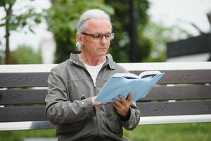 Handsome grandfather sits on a bench in the park and reads a newspaper. Senior gray-haired man. photo