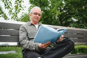 Old man with gray hair reads a book on a bench in the park. Rest in the park. photo