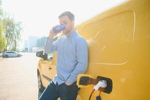 hermoso hombre Bebiendo café mientras cargando eléctrico coche foto