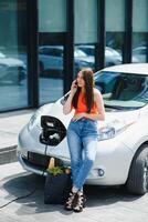 utilizando teléfono inteligente mientras espera. mujer en el eléctrico carros cargar estación a tiempo de día. marca nuevo vehículo. foto