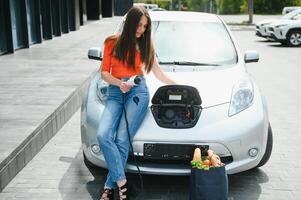 joven mujer es en pie cerca el eléctrico coche. el alquiler coche es cargando a el cargando estación para eléctrico vehículos coche intercambio. foto