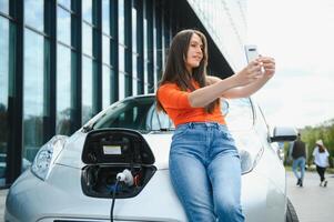 utilizando teléfono inteligente mientras espera. mujer en el eléctrico carros cargar estación a tiempo de día. marca nuevo vehículo. foto