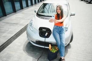 utilizando teléfono inteligente mientras espera. mujer en el eléctrico carros cargar estación a tiempo de día. marca nuevo vehículo. foto