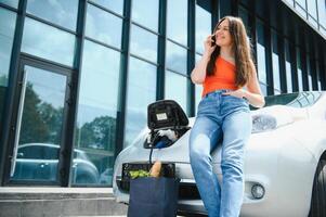 Young woman is standing near the electric car and looks at the smart phone. The rental car is charging at the charging station for electric vehicles. Car sharing. photo