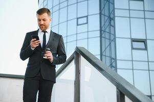 barbado empresario en formal traje en descanso utilizando móvil teléfono utilizar teléfono inteligente negocio hombre en pie fuera de en moderno urbano ciudad calle antecedentes con café taza en céntrico al aire libre. Copiar espacio foto