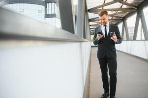 barbado empresario en formal traje en descanso utilizando móvil teléfono utilizar teléfono inteligente negocio hombre en pie fuera de en moderno urbano ciudad calle antecedentes con café taza en céntrico al aire libre. Copiar espacio foto