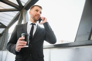 barbado empresario en formal traje en descanso utilizando móvil teléfono utilizar teléfono inteligente negocio hombre en pie fuera de en moderno urbano ciudad calle antecedentes con café taza en céntrico al aire libre. Copiar espacio foto