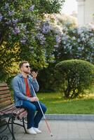 joven ciego hombre con teléfono inteligente sentado en banco en parque en ciudad, vocación. foto