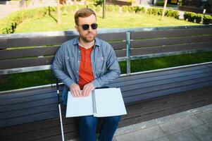 Visually impaired man with walking stick, sitting on bench in city park. Copy space photo