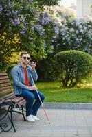 Visually impaired man with walking stick, sitting on bench in city park. Copy space photo