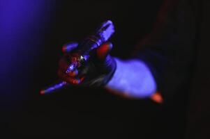 Closeup of tattooer master's hand in black glove holding machine for making tattoo art on body isolated on dark background in neon. photo