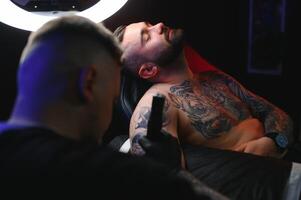 masculino tatuaje artista participación un tatuaje pistola, demostración un proceso de haciendo tatuajes en un masculino tatuado del modelo brazo foto