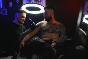 bearded tattoo artist working at his studio tattooing sleeve on the arm of his male client. Man getting tattooed by professional tattooist photo