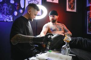 bearded tattoo artist working at his studio tattooing sleeve on the arm of his male client. Man getting tattooed by professional tattooist photo