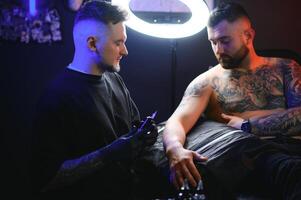 tatuaje artista demuestra el proceso de consiguiendo negro tatuaje con pintar. Maestro trabajos en negro estéril guantes foto