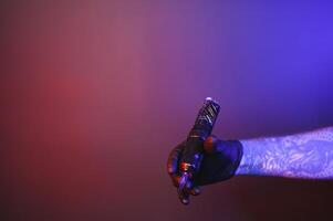 Closeup of tattooer master's hand in black glove holding machine for making tattoo art on body isolated on dark background in neon. photo