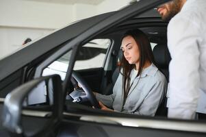 Satisfied happy caucasian female client customer woman sitting at the wheel of new car while male shop assistant helping her choose it photo