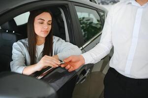 Dealer giving key to cheerful female driver while selling modern red vehicle in dealership photo