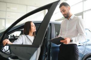 Satisfied happy caucasian female client customer woman sitting at the wheel of new car while male shop assistant helping her choose it photo