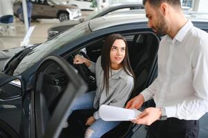 Satisfied happy caucasian female client customer woman sitting at the wheel of new car while male shop assistant helping her choose it photo