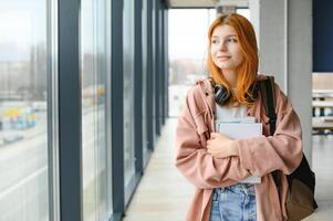 Red-haired student girl stands by the window with books and a backpack photo