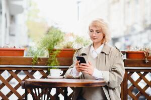 young stylish beautiful woman sitting in city cafe in street, spring fashion trend style, drinking latte, pink purse, exited, emotional, happy, smiling photo