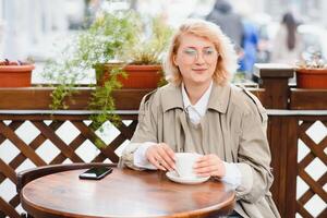 young stylish beautiful woman sitting in city cafe in street, spring fashion trend style, drinking latte, pink purse, exited, emotional, happy, smiling photo